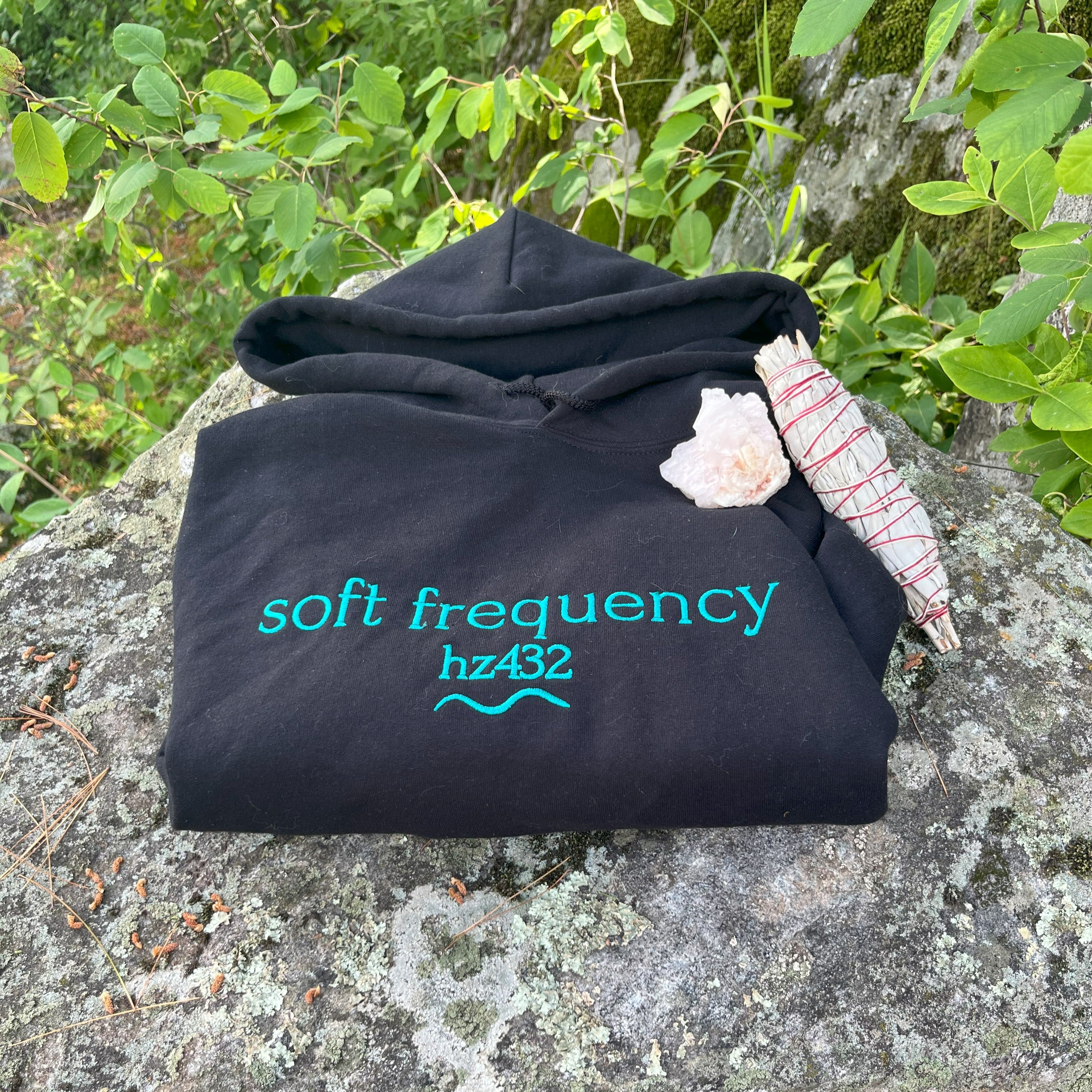 Folded black “soft frequency hz432” hoodie on mossy rock with leaves and clear quartz — intentional fashion set at the natural frequency of harmony. Soft Frequencies™ wearable healing apparel.