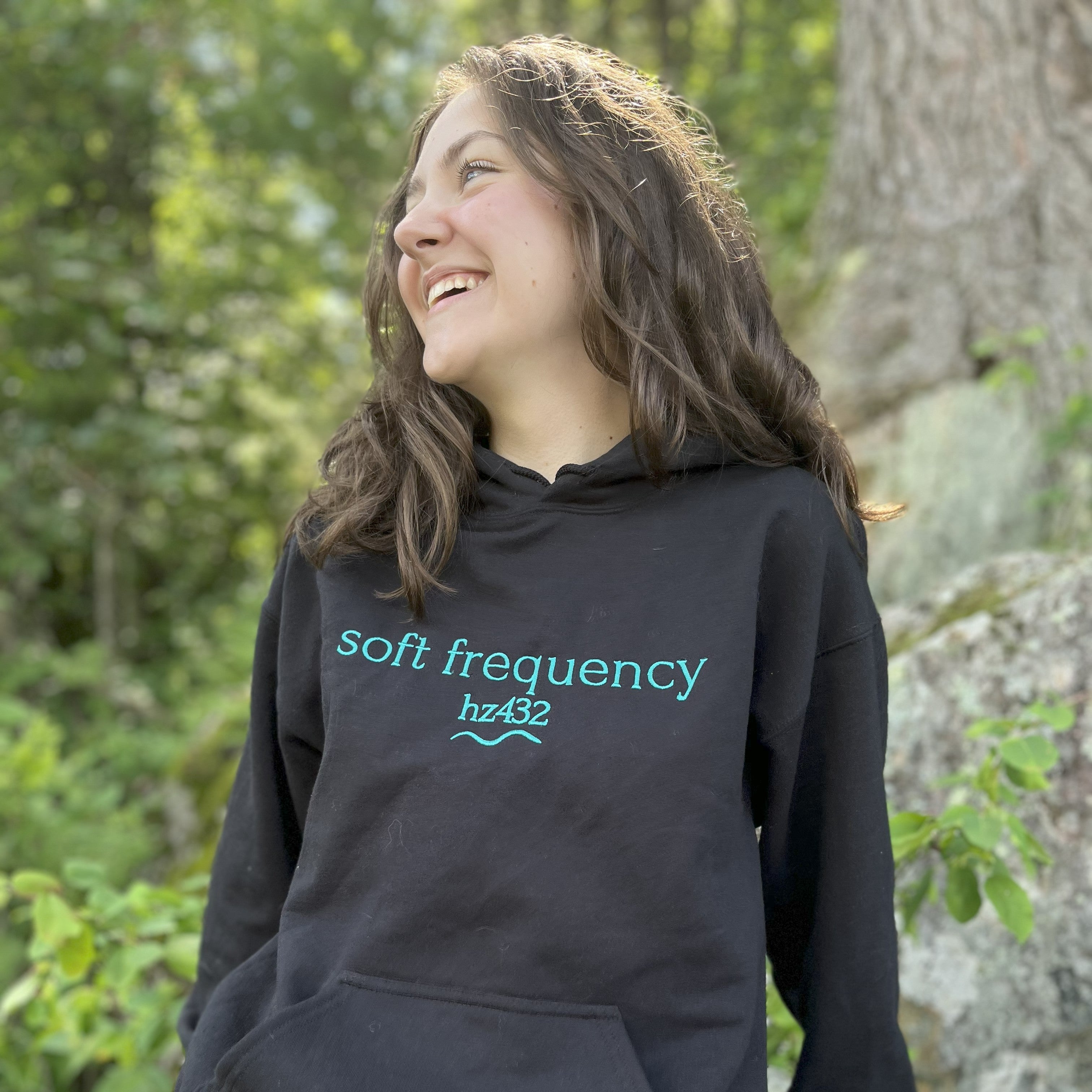 Woman wearing black “soft frequency hz432” hoodie smiling in forest setting — spiritual techwear aligned with 432 Hz. Soft Frequencies™ frequencywear collection.