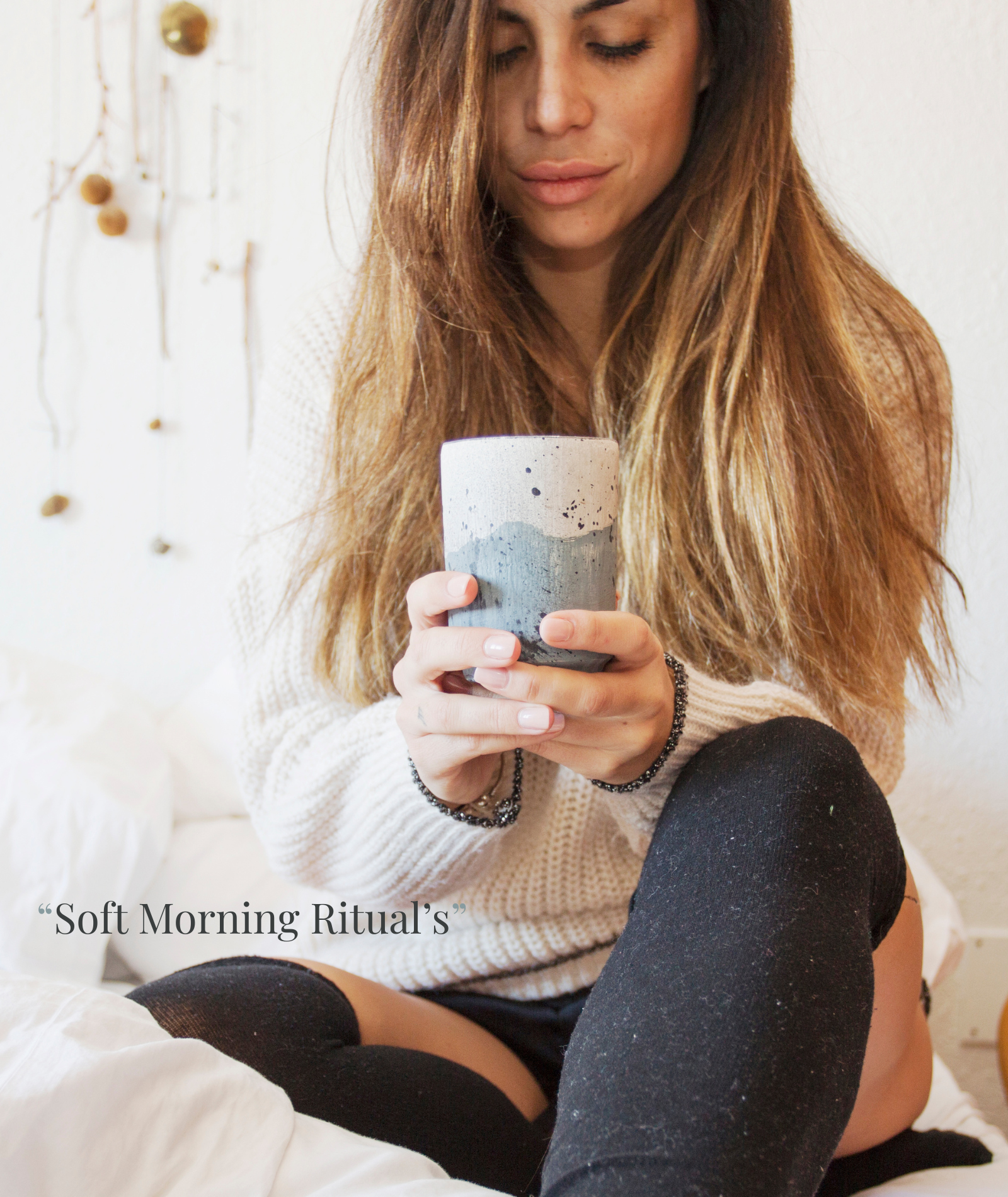 Woman holding ceramic mug on cozy white bedding, wearing sweater, starting her soft morning with intention and calm