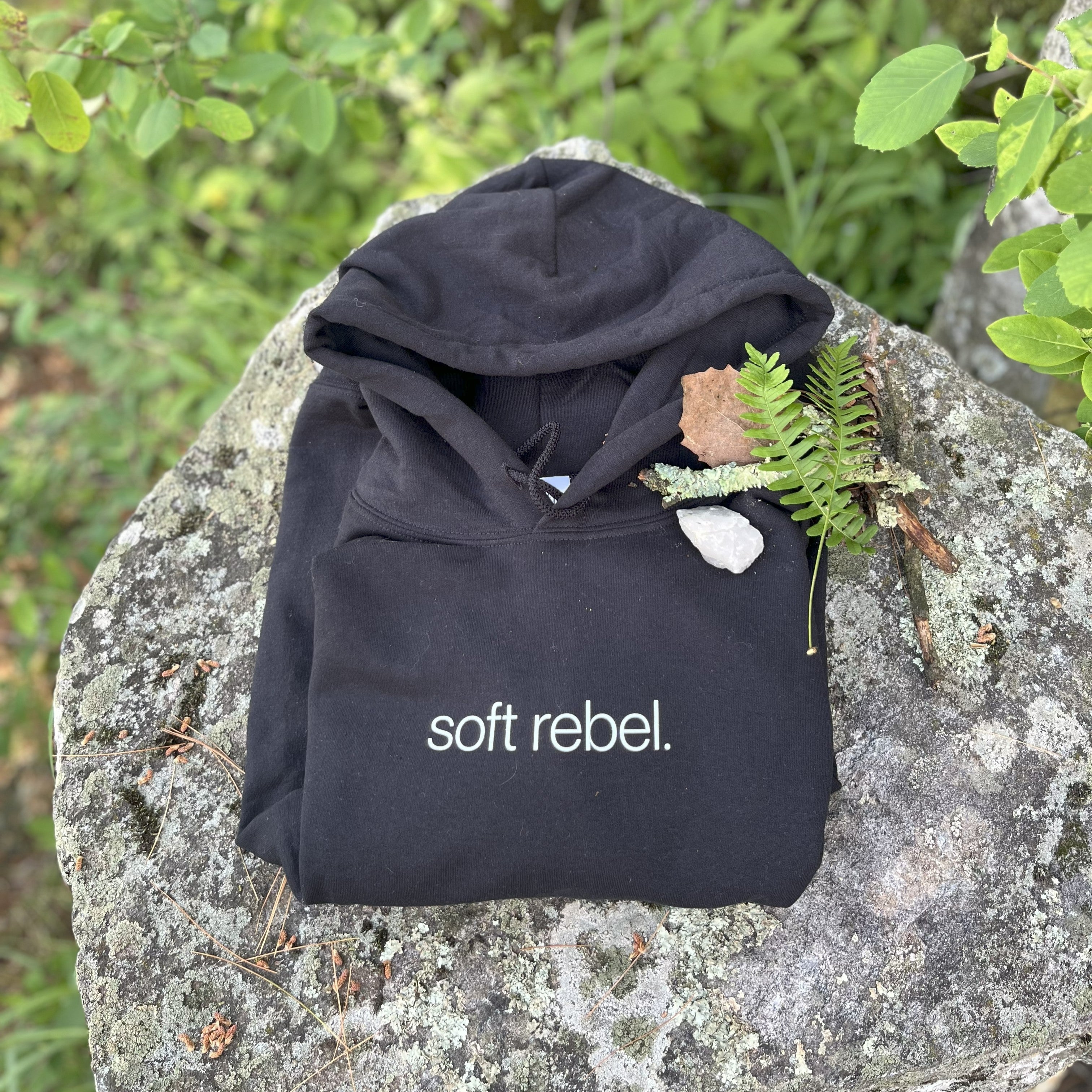 Folded black “soft rebel.” hoodie on mossy stone with clear quartz, dried leaves, and ferns — sacred flat lay of intentional frequencywear by Soft Frequencies™. Minimalist design set at Hz 432.
