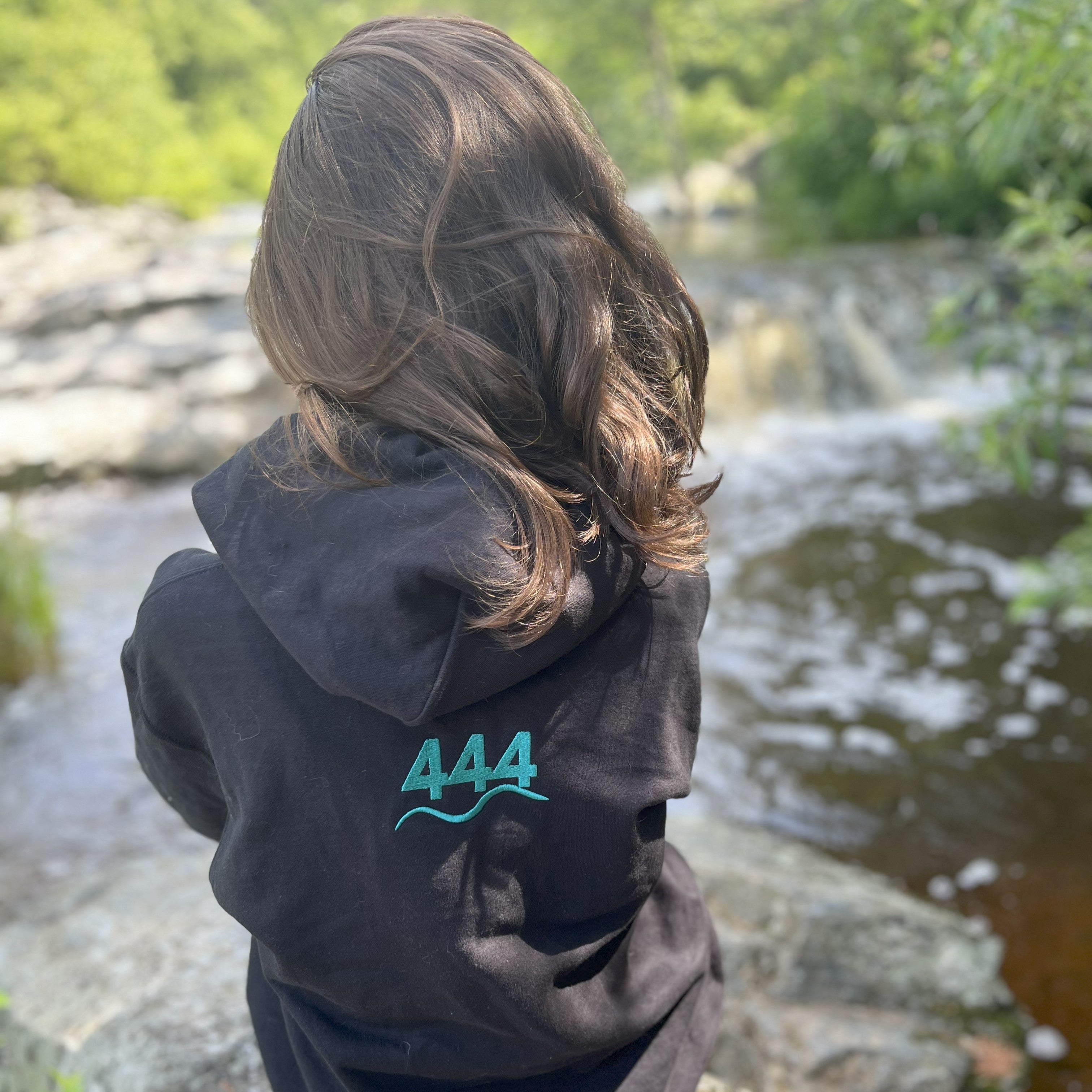 Back view of black hoodie with angel number 444 in teal embroidery — worn by woman near riverbank. Soft Frequencies™ sacred hoodie for spiritual alignment and synchronicity.
