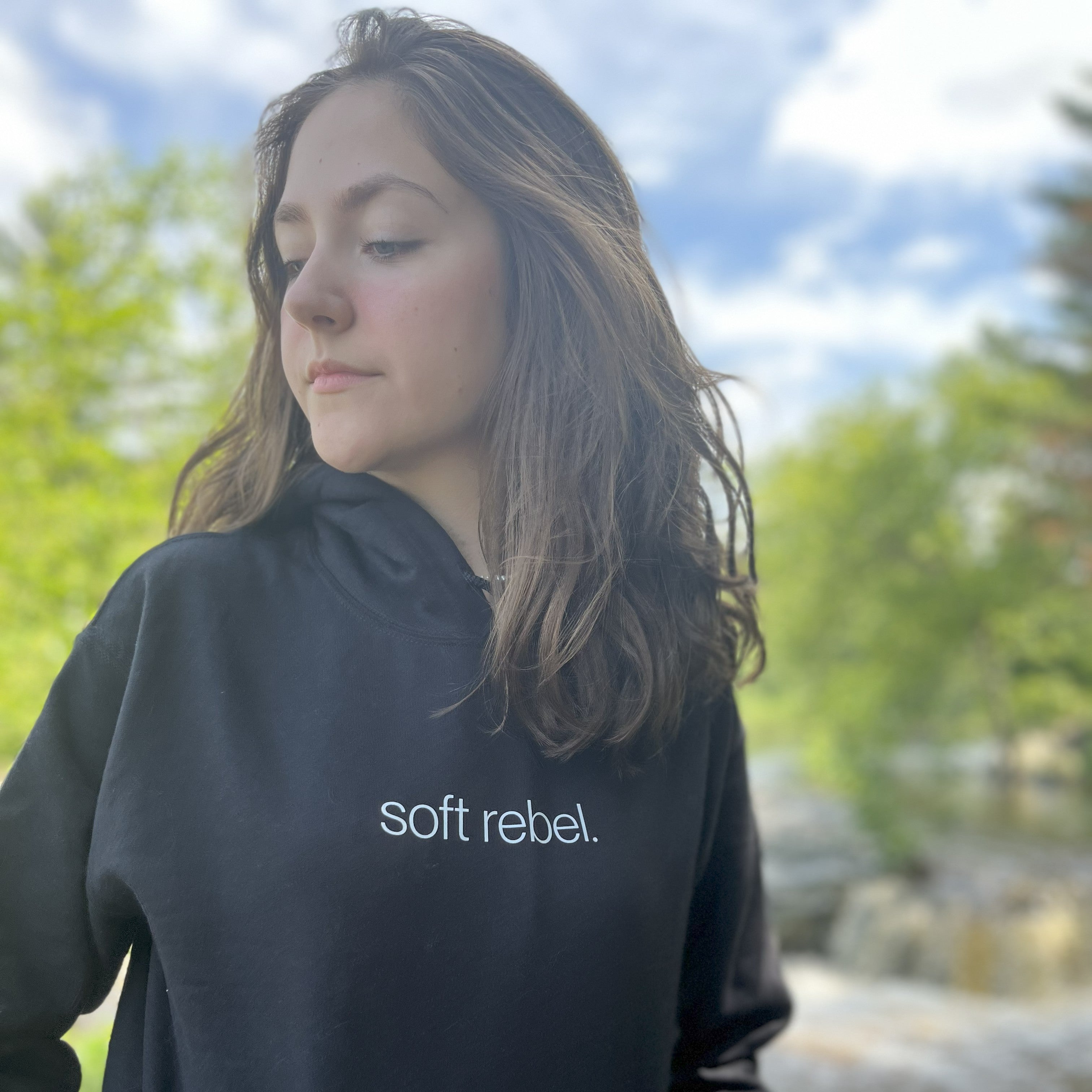 Young woman wearing black “soft rebel.” hoodie standing near forest waterfall, looking serene and empowered — spiritual techwear for mindful rebels. Soft Frequencies™ 432 Hz frequencywear.
